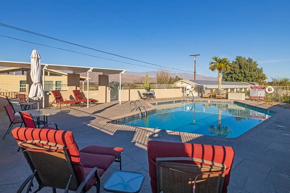 Outdoor motel pool on a sunny desert day, surrounded by red lounge chairs, a pergola, palm tree, and dry mountains in the background